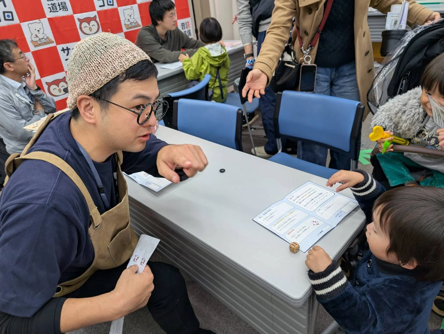 丸メガネ・ニット帽・エプロン姿で、子どもと教材を囲む前田大介